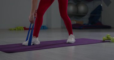 Kırmızı taytlı ve beyaz ayakkabılı bir kız iyi donanımlı bir fitness stüdyosunda bel egzersizine odaklanmış lastik çekme egzersizi yapıyor..