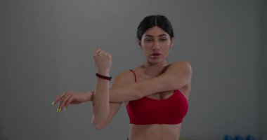 Bir spor salonundaki kız kırmızı spor bluzu giyerken omuz ve kol kaslarını esnetiyor..