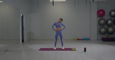 İyi aydınlatılmış bir spor salonunda atletik giyimli bir kız başını eğiyor esnekliğini arttırıyor ve antrenmanına hazırlanıyor. Çevre sağlık ekipmanlarıyla dolu..