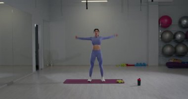 Aynalarla dolu geniş bir spor salonunda atletik kız vücudunu ısıtmak için kollarını ileri geri sallayarak enerjik bir egzersiz yapıyor..