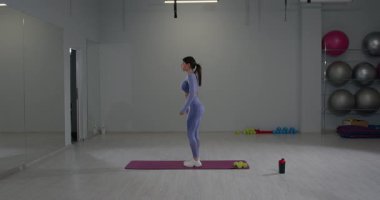 Sportif kız parlak bir fitness stüdyosunda diz esneme hareketleri yapıyor. Yoga minderinin üzerinde duruyor ve ağırlıklarla tekniğine odaklanıyor. Bu aktivite esnekliği ve gücü teşvik eder..