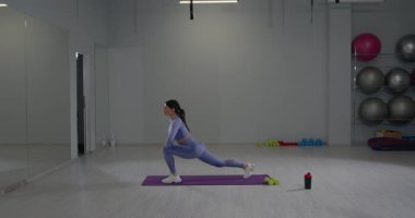 Atletik kız iyi aydınlatılmış bir spor salonunda esnemek için ileri hamleler yapıyor. Spor aletleri ve spor paspaslarıyla çevrili ve kararlı görünüyor..