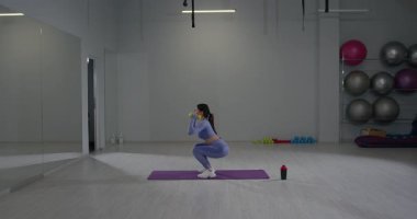 Geniş bir spor salonunda sportif bir kız dambıllara basarken çömelmeye odaklanır. Formunu ve kararlılığını spor aletleriyle sarmaladığı egzersizinde gösteriyor..