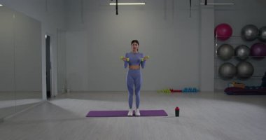 Sportif kız modern bir spor salonunda duruyor ve kendine güvenerek dambıllarla kol egzersizleri yapıyor. Parlak alan spor aletleri ve fitness aksesuarlarını içeriyor.