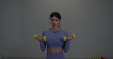 Sportif kız, modern bir spor salonunda kasları üzerinde çalışmak için parlak yeşil dambılları kaldırıyor. Atmosfer enerjiktir ve egzersiz rutinini geliştirmeye odaklanmıştır..
