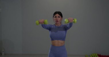 Dişi sporcu hafif dambıllar kullanarak omuz egzersizleri yapar. Öğlen saatlerinde modern bir spor salonunda antrenman yaparken odaklanmış ifadesi kararlılığını gösteriyor..