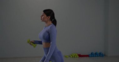 Sportif kız çevikliğini ve gücünü sergileyerek iyi aydınlatılmış bir spor salonunda Bulgar hamlelerini gerçekleştiriyor. Ağırlıklar arka planda çeşitli renklerde görülebilir.