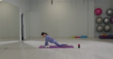 Atletik kız, iyi aydınlatılmış bir spor salonunda yoga minderinin üzerinde ileri doğru hamlesini yapıyor. Egzersiz ekipmanlarıyla çevrili bir antrenmana hazırlanırken esneklik ve denge üzerine odaklanır..