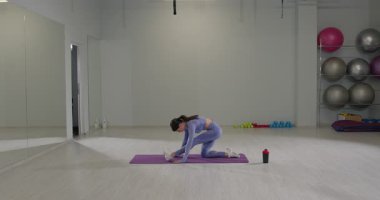 Sportif bir kız, geniş bir spor salonundaki yoga minderinde sırt üstü hareketlerini sergiliyor. Alan egzersiz topları ve ağırlıklarla dolu. Bu da canlı bir egzersiz atmosferi yaratıyor..