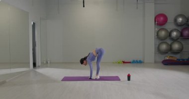 Kız geniş bir spor salonunda esneyebilirliğini göstererek ve sağlığına odaklanarak derin bir viraja giriyor. Bölge iyi ışıklandırılmış ve yakınlarda egzersiz ekipmanları var..