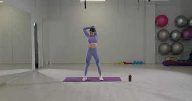 Sportif kız iyi aydınlatılmış bir spor salonunda triseps oynuyor. Mor bir spor kıyafeti giyiyor ve arka planda spor malzemeleri olan bir paspasın üzerinde duruyor..