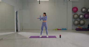 Sportif kız iyi aydınlatılmış bir spor salonunda omuzlarını esnetiyor. Aktif bir antrenman seansına hazırlanırken odaklanmış görünüyor. Spor salonu egzersiz topları ve ağırlıklarla donatılmıştır..