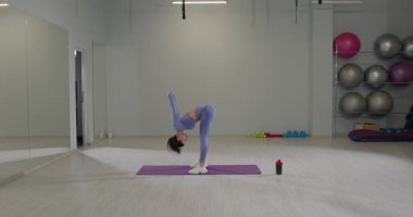 Bir spor salonunun içinde mor elbiseli bir kız yoga minderinde denge kurarken kollarını geriye doğru uzatıyor. Atmosfer parlak ve egzersiz için motive edici..