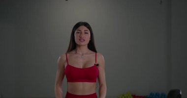 Sportif kız spiker, fitness stüdyosu ortamında kameraya güvenle konuşuyor. Kırmızı bir kıyafet giyip spor bahşişlerini paylaşmaya ya da sağlıklı bir yaşam tarzı geliştirmeye hazır..
