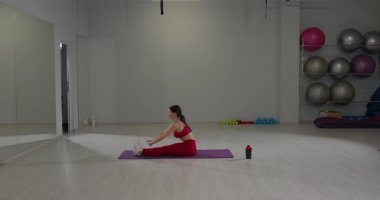 Kız, iyi aydınlatılmış bir fitness stüdyosunda sırtına ve bacaklarına odaklanarak esnekliğini artırmak için oturarak esneme hareketi yapıyor. Egzersiz ekipmanları arka planda görünür..