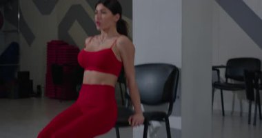 Modern bir spor salonunda kırmızı etek giyen bir fitness meraklısı ters sandalye şınavı çeker. Bu egzersiz üst gövde dayanıklılığını ve istikrarını artırırken trisepsleri hedef alır..