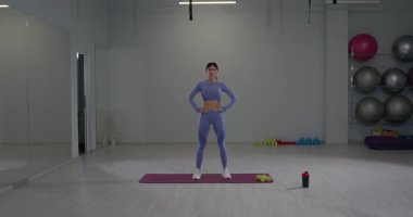 Atletik kız, iyi donanımlı bir spor salonunda ısınma rutininin bir parçası olarak sağa sola dönüyor. Çevrede çeşitli fitness ekipmanları ve spor paspası bulunmaktadır..