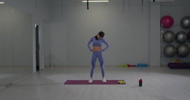 Parlak bir fitness stüdyosunda sportif bir kız antrenmanına odaklanır ve sağa sola kafa sallar. Atletizm kıyafeti giyiyor ve formda olduğunu gösteriyor..