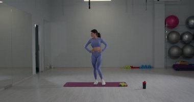 Aktif kıyafet giyen genç bir kadın egzersiz rutinine hazırlanmak için dizlerini esnetiyor. Geniş fitness stüdyosu onun odaklanmasını ve motivasyonunu arttırıyor..
