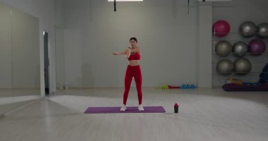 Çağdaş fitness stüdyosundaki kırmızı spor elbiseli kız omuzlarını ve trisepslerini esneterek egzersiz rutininin bir parçası olarak esneklik ve kas gevşemesine odaklanıyor..