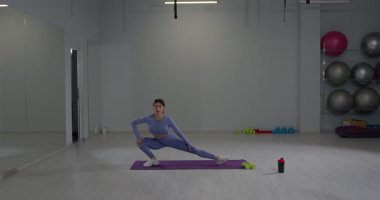 Sporcu kız, geniş bir spor salonunun içinde sağa sola hamlelerle meşgul. Çevre, arka planda görülebilen egzersiz ekipmanlarıyla aydınlandı..