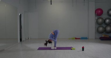 Atletik kız, geniş bir spor salonundaki yoga minderinde öne eğilerek esniyor. Ayar parlak ve iyi donanımlı ağırlıklar ve egzersiz topları içeriyor..