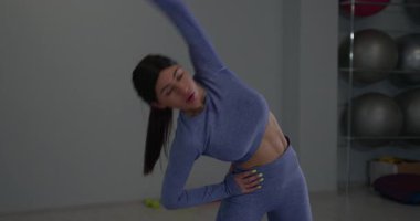 Parlak bir fitness stüdyosunda atletik bir kız sağa sola eğiliyor. Dengeyi korumaya odaklanması, uygunluk ve esnekliğe olan bağlılığını vurguluyor..