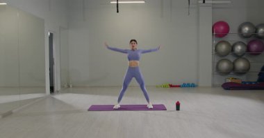 Mor kıyafetli atletik kız, iyi aydınlatılmış bir spor salonundaki yoga minderinin önünde kıvrılıyor. Egzersiz ekipmanları görülebiliyor..