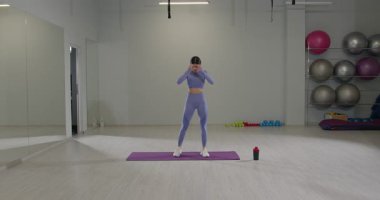 Sportif kız, geniş bir spor salonunda yoga minderinin üzerinde duruyor ve yoğun bir antrenmana hazırlanırken saç bandını ayarlıyor. Doğal ışık odayı dolduruyor ve motivasyon atmosferini arttırıyor..