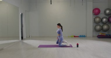 Kendini işine adamış bir kız parlak bir spor salonunda ileri atlama hareketini gerçekleştiriyor. Oda egzersiz paspasları ve spor yapmak için ideal spor ekipmanları ile donatılmıştır..