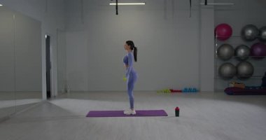 Sportif kız, modern bir spor salonunda spor minderinin üzerinde duruyor ve dambıllarla kol egzersizleri yapıyor. Mekan spor aletleri ve aynalarla dolu. Enerjik bir atmosfer yaratıyorlar..
