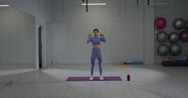 Sportif kız, iyi aydınlatılmış bir spor salonunda, kafasının üstünde dambıllarla çömelme egzersizleri yapıyor. Mekanda egzersiz ekipmanları ve paspaslar yer alıyor.