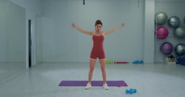 Kız sporcu bir fitness stüdyosunda paspasın üzerinde dururken düz kollarıyla rotasyon egzersizi yapıyor. Çevre aydınlıktır ve çalışma ekipmanlarına sahiptir..