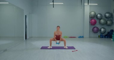 Genç sporcu kız, geniş ve iyi donanımlı bir fitness stüdyosunda ağırlık tutarken sumo çömelmesi yapıyor. Çevre, dayanıklılık eğitimi ve uygunluğa odaklanmayı yansıtıyor.