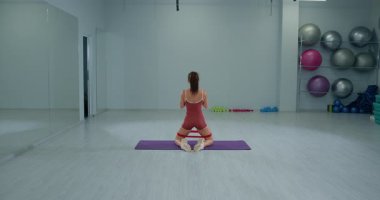 Kız sporcu, parlak bir fitness stüdyosunda dizlerinin üstüne çöküp çömeliyor. Oda egzersiz topları ve ağırlıklarla dolu. Enerjik bir atmosfer yaratıyor..