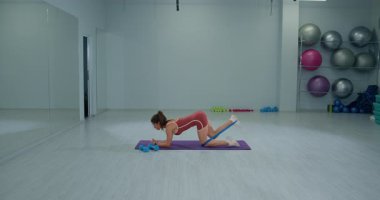 Kız sporcu, iyi aydınlatılmış bir spor salonunda eşşek tekmelerine odaklanmış bir fitness rutiniyle meşgul. Formunu korurken aynı zamanda egzersiz etkinliğini arttırmak için bandı kullanıyor..