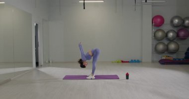 Mor elbiseli kız yoga stüdyosunda esneme hareketleri yaparken öne eğiliyor. Geniş bir alanda egzersiz ekipmanları ve paspaslarla çevrili ve odaklanmış durumda..
