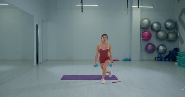Kız sporcu geniş bir spor salonunda dambılları tutarken ters hamleler yapıyor. Çevrede egzersiz ekipmanları ve etkin bir fitness rutinini gösteren bir spor minderi bulunuyor..