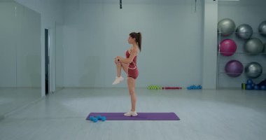 İyi aydınlatılmış bir spor salonunda diz sarılma egzersizi yapan esnek ve güçlü bir kız sporcu. Arka planda çeşitli fitness ekipmanları görünür.