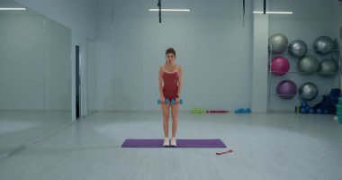 Genç bayan sporcu, iyi aydınlatılmış bir spor salonundaki bir paspasın üzerinde düz bir dambıl kaldırışı gerçekleştiriyor. Atmosfer, güç eğitimine odaklanmış durumda..