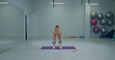 Kız sporcu geniş bir duruş sergiliyor. Parlak bir fitness stüdyosunda dambıllarla Romen kaldırışı yapıyor..