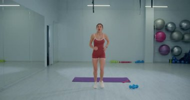 Girl athlete is performing butt kicks on a gym mat in a well lit fitness studio. She is wearing a red workout outfit and focuses on improving her agility and warm up routine.