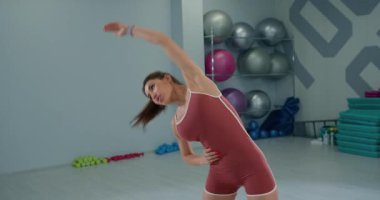 Geniş bir fitness stüdyosunda bir kız sporcu, esneme hareketleri yaparken kolunu sağa sola eğiyor. Harekete odaklanması esneklik ve güç eğitimini vurguluyor.