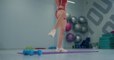 Genç bayan sporcu modern bir spor salonunda ayakta duran bir diz kaldırma egzersizi yapıyor. Spor salonunda spor aletleri ve canlı renkler yer alıyor. Güç ve denge üzerine odaklanır..