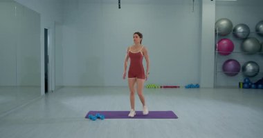 Kız sporcu bir dizini dizine dayayıp diz sarılma egzersizi yapıyor. Çeşitli fitness ekipmanlarıyla donatılmış iyi aydınlatılmış bir spor salonunda pratik yapıyor..