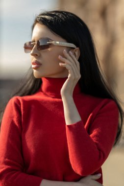 Kırmızı kazaklı güzel bir kız, güneşli bir günde açık havada eğlenirken kendine güvenen gözlüklü. Düşünceli duruşu zarafet ve stili yansıtıyor.