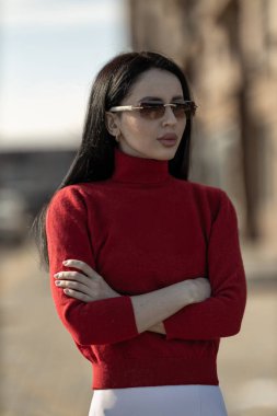 Gözlüklü güzel kız, kollarını kavuştururken canlı kırmızı bir kazak giyiyor.