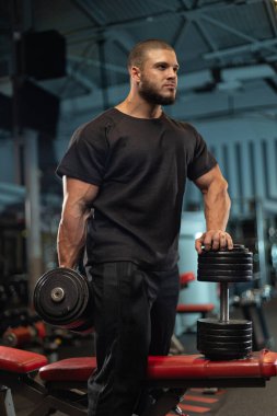 Erkek sporcu, iyi donanımlı spor salonu atmosferinde güç antrenmanına hazırlanırken güven içinde ayakta duruyor. Kararlılık ve odaklanma dolu.