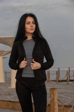 Kadın, su kenarındaki ahşap bir güvertede, siyah atletik giysiler içinde duruyor. Gün batımının yumuşak renkleri etrafında huzurlu bir atmosfer yaratıyor.