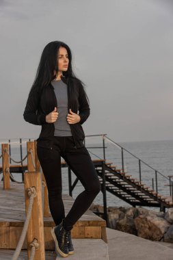 Siyah ceketli ve kot pantolonlu bir kadın güvenli bir şekilde tahta iskelede duruyor, yumuşak bir akşam gökyüzünün altındaki dingin denizi seyrediyor atmosfer huzurlu ve yansıtıcı.